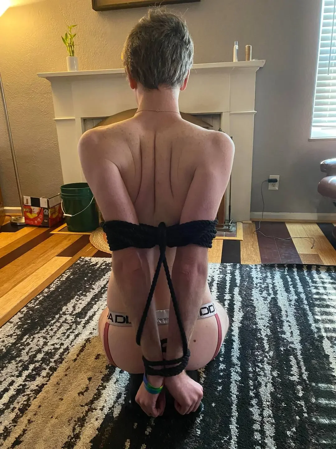 indoors, ass, shaved, nude, solo, partially dressed, sitting, panties, male, from behind, male only, 1boy, on floor, white male, underwear, jewelry, short hair, bdsm, back, bondage, arms behind back, bound, bound wrists, facing away, median furrow, rug