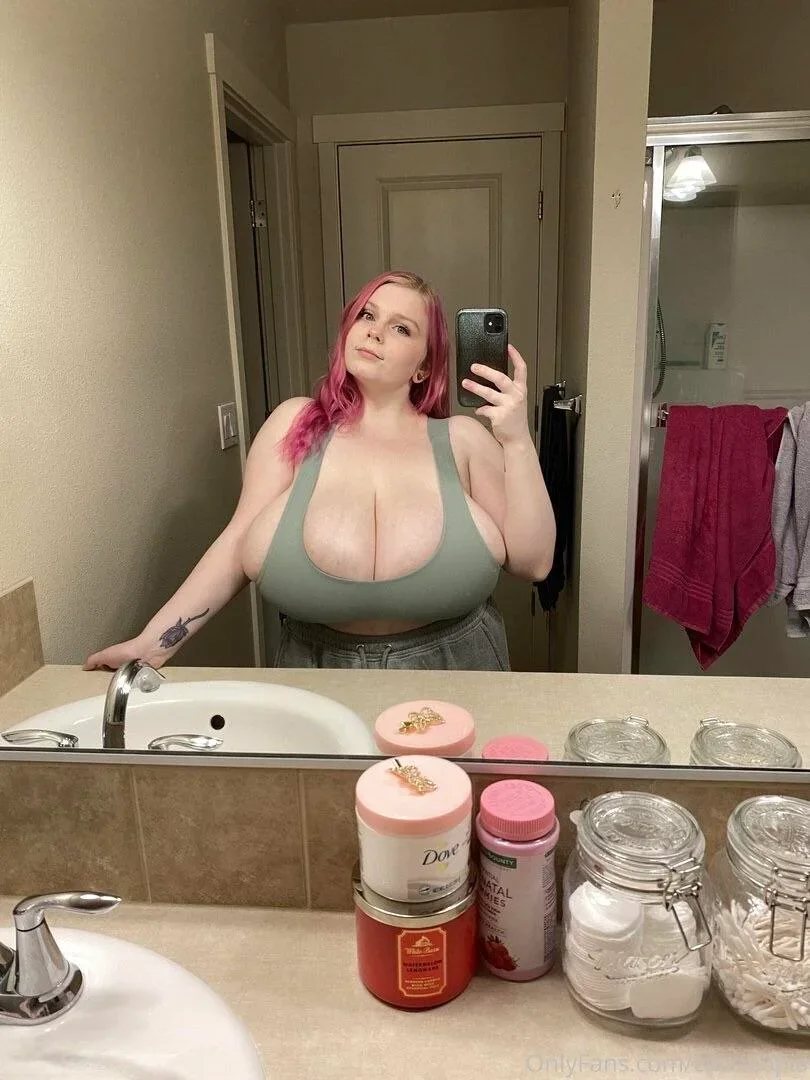 cassie0pia, 1girl, female only, white female, looking at viewer, long hair, front view, standing, pale-skinned female, selfie, smile, dyed hair, non-nude, tattoo, bra, mirror selfie, blue eyes, bathroom, cleavage, huge breasts, sleeveless, pink hair, tank top, overflowing breasts