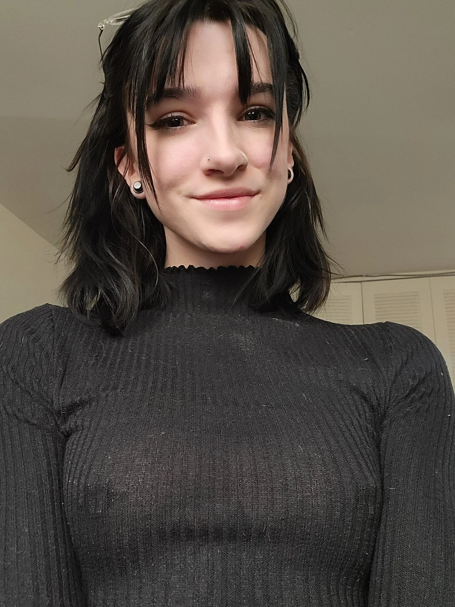 motzie, motziee, 1girl, female only, white female, breasts, looking at viewer, front view, medium hair, standing, pale-skinned female, selfie, black hair, piercing, smile, fringe, bangs, non-nude, nose piercing, black clothes, no bra, wavy hair, ear piercing, american female, nipple bulge, sweater, flesh tunnel, glasses on head